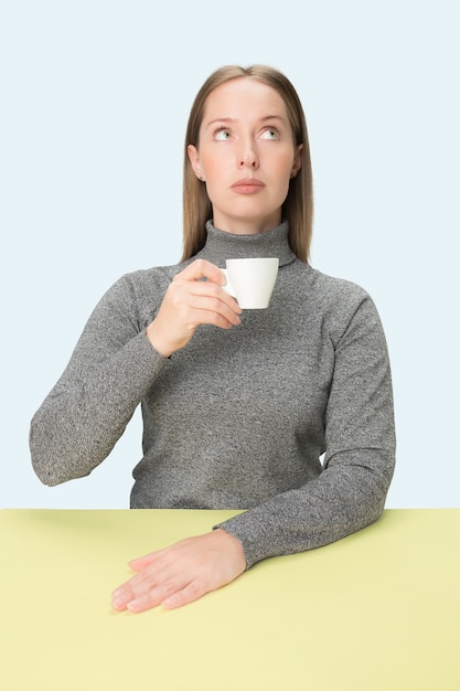 Mooie eenzame vrouw zit op blauwe studio en kijkt verdrietig met het kopje koffie in de hand.