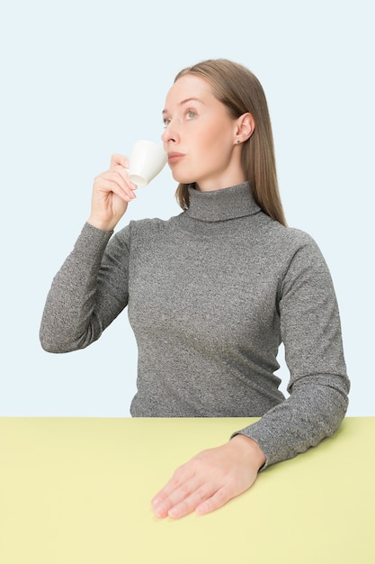 Mooie eenzame vrouw zit op blauwe studio en kijkt verdrietig met het kopje koffie in de hand.