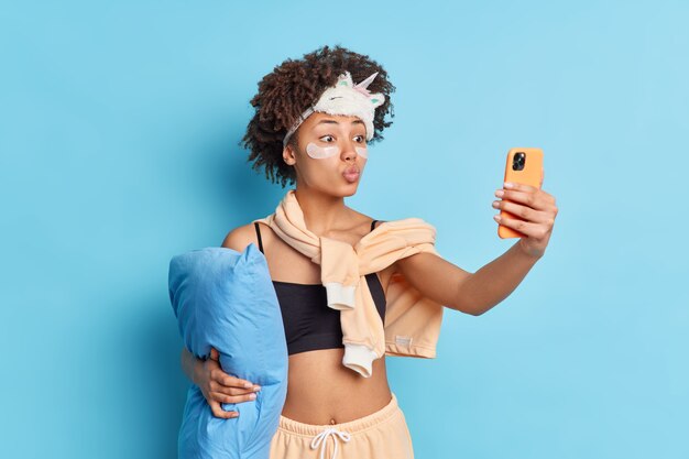 Mooie etnische vrouw maakt foto van zichzelf houdt lippen afgerond houdt smartphone vooraan poses in casual binnenlandse pyjama slaapmasker op voorhoofd geïsoleerd op blauwe achtergrond. Selfie voor het slapengaan