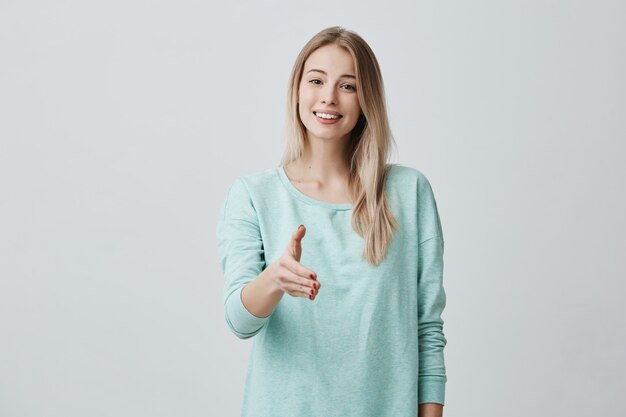 Mooie Europese vriendelijke vrouw met lang blond haar, gekleed in een casual blauwe trui die breed lacht en haar witte perfecte tanden demonstreert en haar arm strekt tijdens de introductie. Lichaamstaal