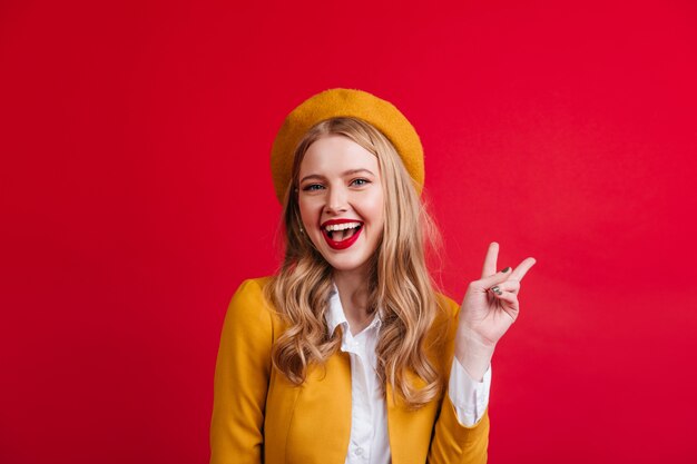 Mooie Franse jonge vrouw vredesteken tonen. Vooraanzicht van aantrekkelijk blondemeisje dat op rode muur wordt geïsoleerd.