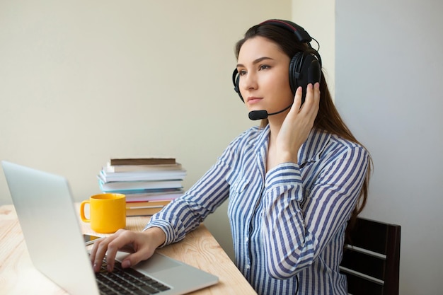 Gratis foto mooie freelance operator die werkt in online marketing met headsets en een laptop op een desktop op kantoor. vrolijke callcentervrouw die vanuit huis werkt en met de klant praat