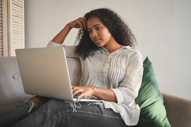 Mooie geconcentreerde jonge donkere vrouw met Afro-kapsel studeert op afstand via online cursussen, met behulp van wifi op haar laptop terwijl ze thuis op de bank zit. Mensen, technologie en onderwijs