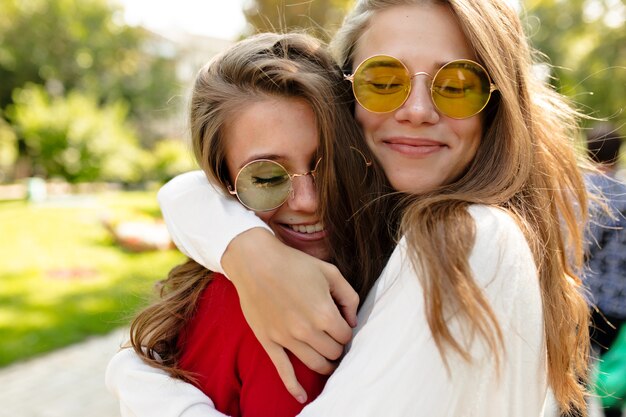 Mooie gelukkige meisjes die buiten in zonnige dag lopen. mooie mooie vrouw in heldere glazen knuffelen haar vriend en gesloten ogen met een grote glimlach, beste vrienden, zussen, posivite stemming