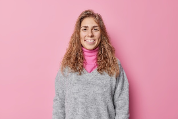 Mooie gelukkige vrouw met lang haar glimlacht graag heeft schone natuurlijke huid ni make-up voelt zich gelukkig draagt casual jumper poses tegen roze achtergrond. Positieve menselijke gezichtsuitdrukkingen en gevoelens