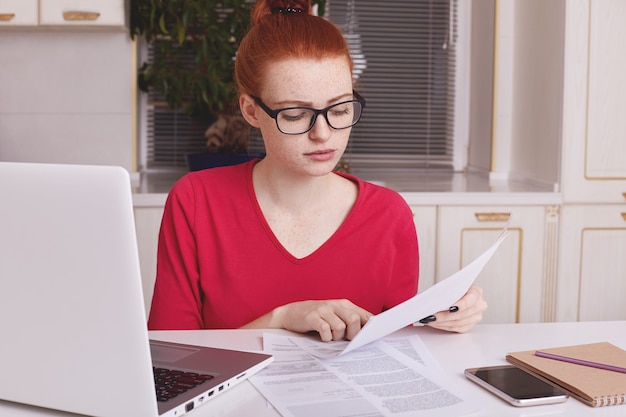 Mooie gember vrouwelijke freelancer werkt ver thuis, bestudeert documenten, zit achter geopende laptop