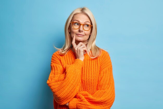 Mooie gerimpelde vrouw van middelbare leeftijd houdt wijsvinger op wang staat in bedachtzame pose dagdromen over iets dat een gebreide trui draagt.