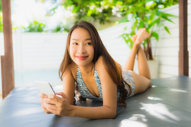 Mooie glimlach van de portret de mooie jonge aziatische vrouw ontspant met mobiele telefoon rond zwembad