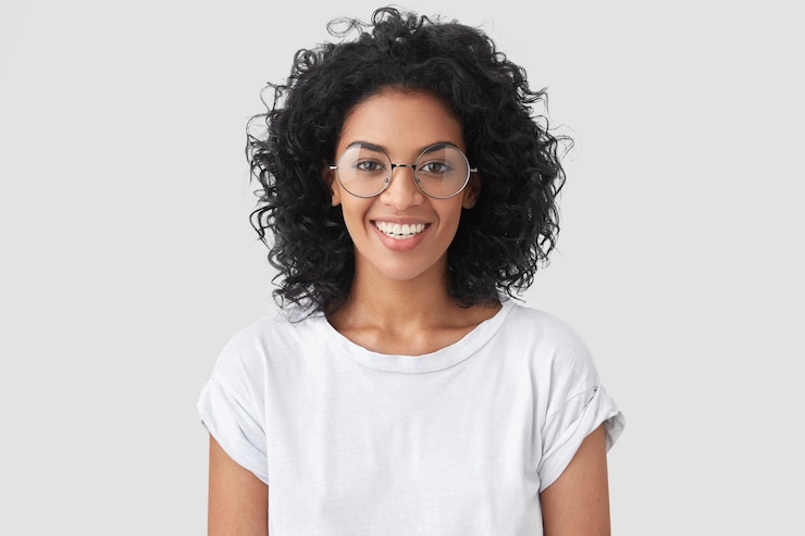 Gratis Foto mooie glimlachende afro-amerikaanse vrouw met knapperig haar, brede glimlach, toont witte tanden, draagt casual t-shirt en bril, staat over muur verheugt zich met vrije dag. vrouw journalist binnen