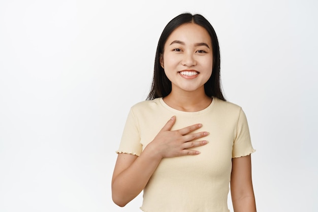 Mooie glimlachende aziatische vrouw houdt de hand op de borst terwijl ze naar zichzelf wijst en er gelukkig uitziet terwijl ze op een witte achtergrond staat