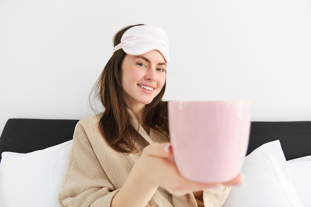 Mooie glimlachende moderne vrouw in pyjama's en slaapmasker die een kopje thee of koffie in bed aanbiedt en