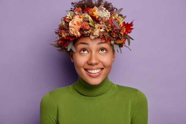 Mooie herfst Afro meisje met blije uitdrukking, kijkt boven op mooie herfst krans gemaakt van seizoensplanten, draagt groene coltrui, geïsoleerd op paarse achtergrond.