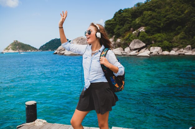 Mooie hipster vrouw reist de wereld rond met rugzak, glimlachend, gelukkig, positief, luisteren muziek in koptelefoon, blauwe tropische oceaan achtergrond, zonnebril, sexy, zomervakantie,