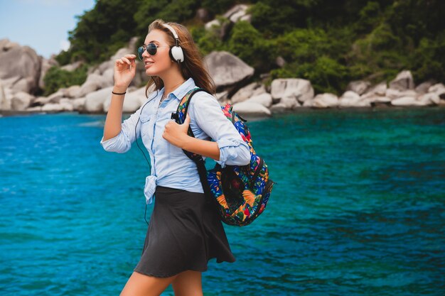 Mooie hipster vrouw reist de wereld rond met rugzak, glimlachend, gelukkig, positief, luisteren muziek in koptelefoon, blauwe tropische oceaan achtergrond, zonnebril, sexy, zomervakantie,