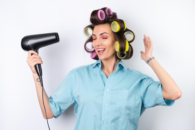 Mooie huisvrouw Jonge vrolijke vrouw met haarkrulspelden en föhn klaar voor een date night maakt een kapsel thuis op een witte achtergrond