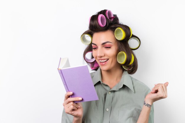 Mooie huisvrouw Jonge vrolijke vrouw met krulspelden lichte make-up met een boek in haar handen op een witte achtergrond Nadenken over een dinerrecept Op zoek naar voedselideeën