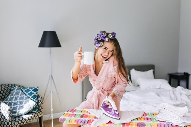 Mooie huisvrouw met lang haar in roze badjas met krultang op hoofd thuis bij het strijken van kleren. Ze sprak aan de telefoon, met een strijkijzer en een beker, glimlachend.