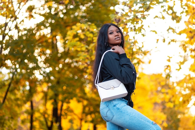 Mooie jonge afro-amerikaanse modelvrouw in modekleding met casual jasje met stijlvolle spijkerbroek en een modieuze handtas in het park tegen een achtergrond van felgeel herfstgebladerte