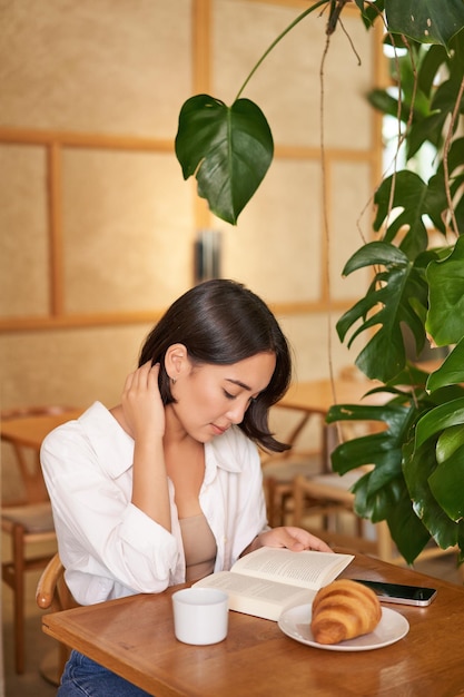 Mooie jonge aziatische vrouw die in café zit met een boek dat een croissant eet en een drinkbeker leest