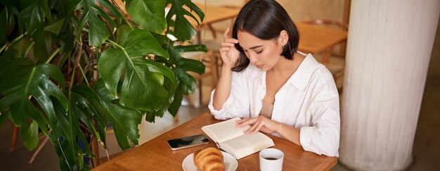 Gratis foto mooie jonge aziatische vrouw die in een café zit met een boek dat een croissant eet en een drinkbeker leest