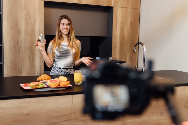 Mooie jonge blonde vrouw blogger drinkwater in de keuken met gezond voedsel op tafel