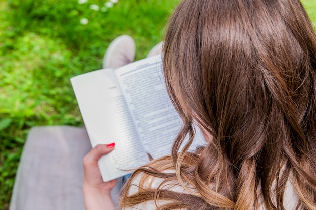 Gratis foto mooie jonge brunette zittend op een verse lente in een park, een boek lezen. close-up van een mooie jonge vrouw lezen boek op park. vrouw geniet van een boek lezen buiten