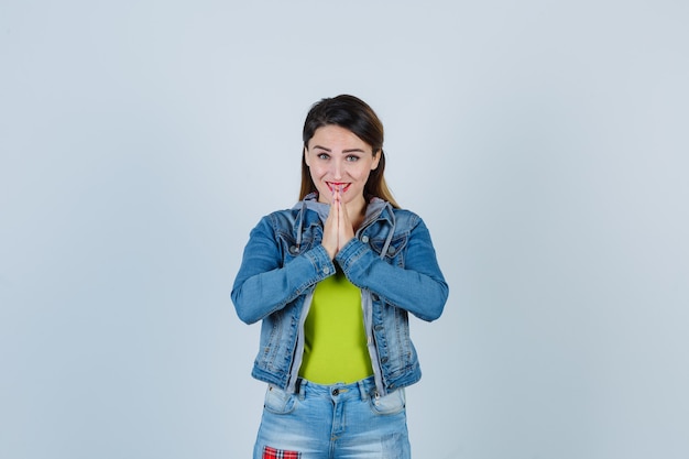 Mooie jonge dame die gevouwen handen toont in een smekend gebaar in denim outfit en er vrolijk uitziet. vooraanzicht.