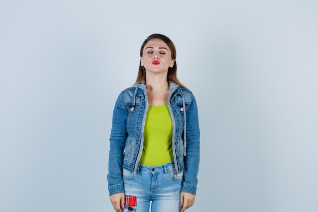 Mooie jonge dame die haar ogen dicht houdt, lippen pruilt in denim outfit en er gracieus uitziet. vooraanzicht.