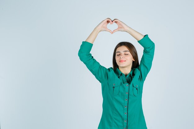 Mooie jonge dame die hartgebaar boven het hoofd in groen overhemd maakt en ontspannen kijkt. vooraanzicht.