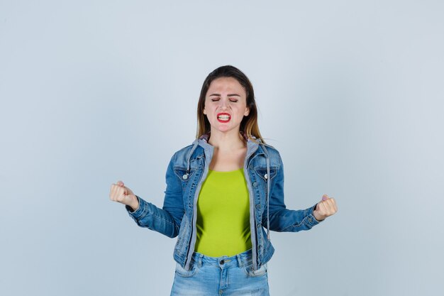 Mooie jonge dame die winnaargebaar toont, ogen sluit, tanden op elkaar klemt in denimoutfit en er gelukkig uitziet. vooraanzicht.