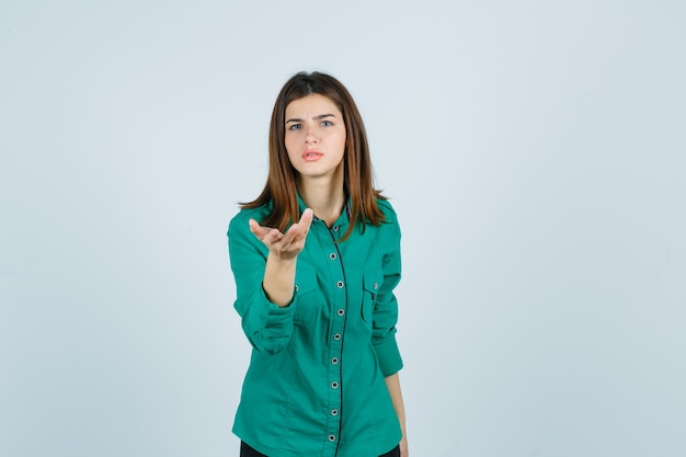 Mooie jonge dame die zich uitstrekt hand in vragend gebaar in groen shirt en op zoek verbaasd, vooraanzicht.