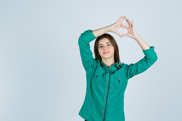 Mooie jonge dame hart gebaar maken in groen shirt en op zoek vrolijk. vooraanzicht.