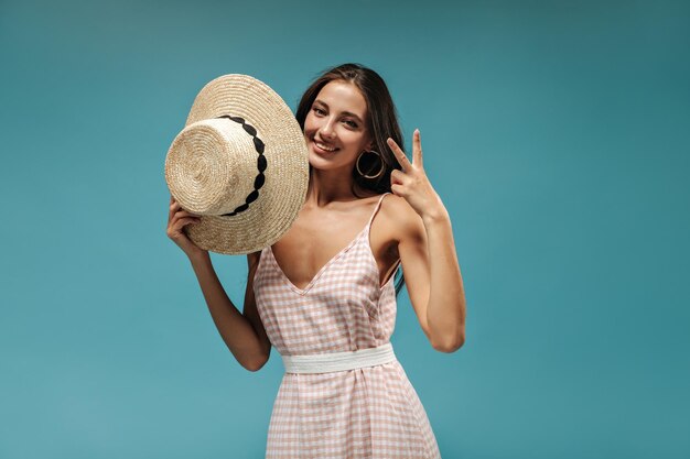 Mooie jonge dame in roze zomerkleren die lacht met vredesteken en moderne hoed vasthoudt op geïsoleerde blauwe achtergrond