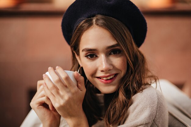 Mooie jonge dame met donkerbruin golvend haar en donkere ogen, trendy make-up, in baret en pullover buiten zitten met een kopje thee en glimlachen