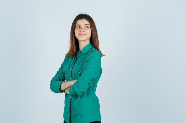 Mooie jonge dame permanent met gekruiste armen terwijl wegkijken in groen shirt en op zoek vrolijk, vooraanzicht.