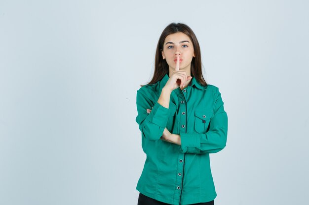 Mooie jonge dame stilte gebaar in groen shirt tonen en voorzichtig kijken. vooraanzicht.