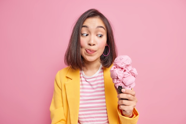 Mooie jonge donkerharige Aziatische vrouw kijkt naar smakelijk lekker ijs likt lippen met tong kan niet wachten tot het eten van een heerlijk zomerdessert gekleed in modieuze kleding. Zoete tand concept