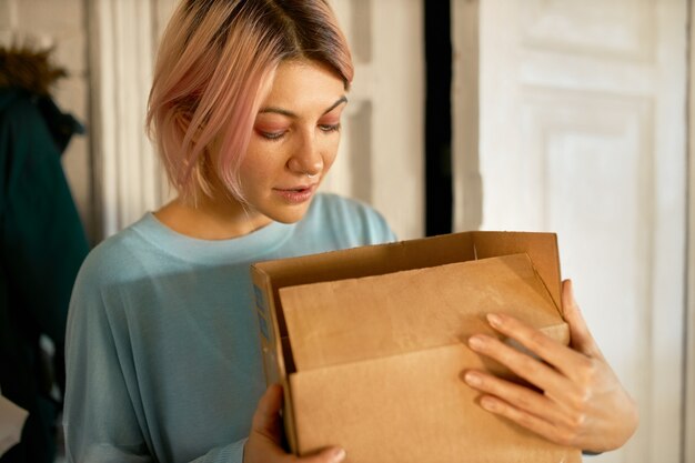 Mooie jonge Europese vrouw poseren binnenshuis met kartonnen doos in haar handen, openen, naar binnen kijken.