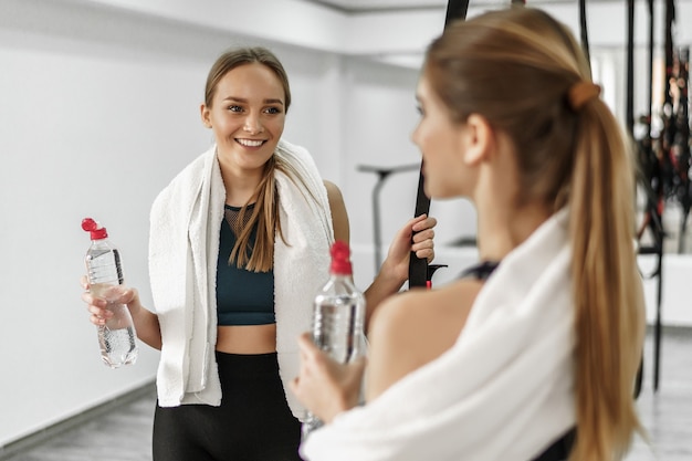 Mooie jonge fitnessmeisjes houden flessen water vast, kijken elkaar aan en glimlachen