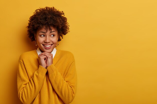 Mooie jonge gekrulde vrouw heeft een vrolijke bui, houdt de handen onder de kin en lacht positief, kijkt opzij, roept prettige herinneringen op, draagt een losse trui, poseert tegen de gele muur, lege ruimte rechts