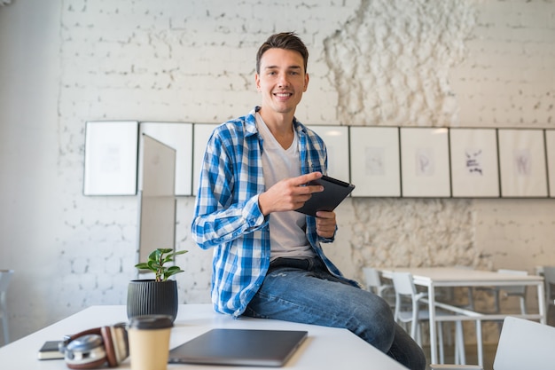 Mooie jonge knappe man in chekered shirt zittend op tafel met behulp van tabletcomputer in co-working office,