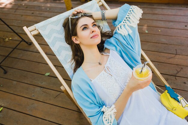 Mooie jonge lachende stijlvolle vrouw zitten in ligstoel in zomer-outfit, het dragen van witte jurk, blauwe cape, zonnebril, portemonnee, vers sap drinken, ontspannen