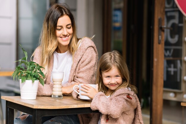 Mooie jonge moeder met mooie dochtertje gekleed in warme truien zitten in cafetaria