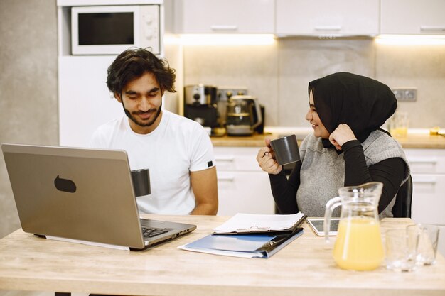 Mooie jonge paar met behulp van een laptop, schrijven in een notitieblok, zittend in een keuken thuis. Arabisch meisje dat zwarte hidjab draagt.