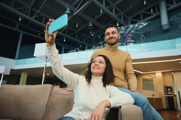 Mooie jonge verliefde jonggehuwden op hun huwelijksreis die een zelfportret selfie maken op een smartphone terwijl ze samen ontspannen in de luchthavenlounge terwijl ze wachten op het instappen van een vlucht.