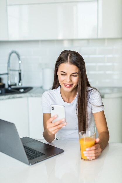 Mooie jonge vrouw die aan laptop gebruikstelefoon werkt in de keuken