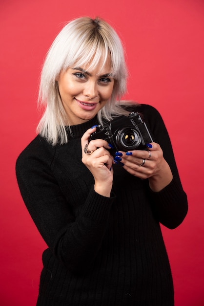 Gratis foto mooie jonge vrouw die een camera houdt terwijl hij tegen rode achtergrond staat. hoge kwaliteit foto