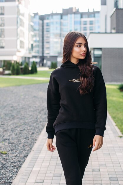 Mooie jonge vrouw die een comfortabel trainingspak draagt terwijl ze langs de straat loopt met gebouwen op de achtergrond. Vrouwelijke mode. Stadslevensstijl