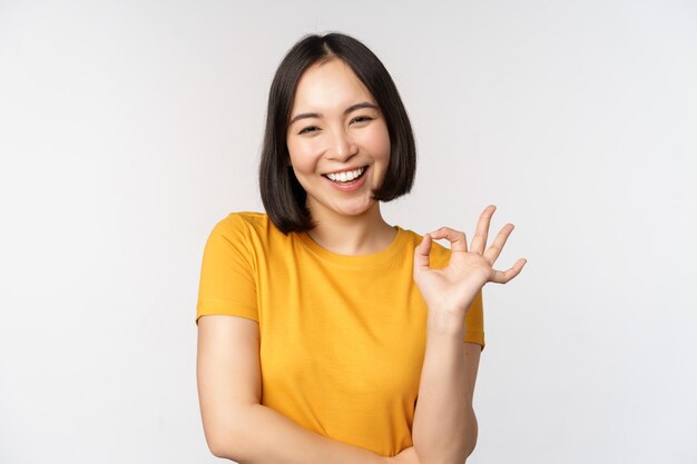 Mooie jonge vrouw die een goed teken toont dat glimlacht en blij aanbeveelt om een soortgelijk product goed te keuren dat in een gele t-shirt staat op een witte achtergrond