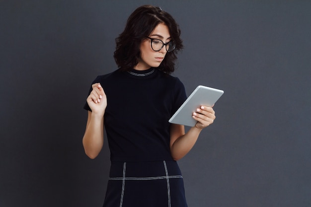 Mooie jonge vrouw die glazen dragen die over grijze muur stellen en tablet in handen houden